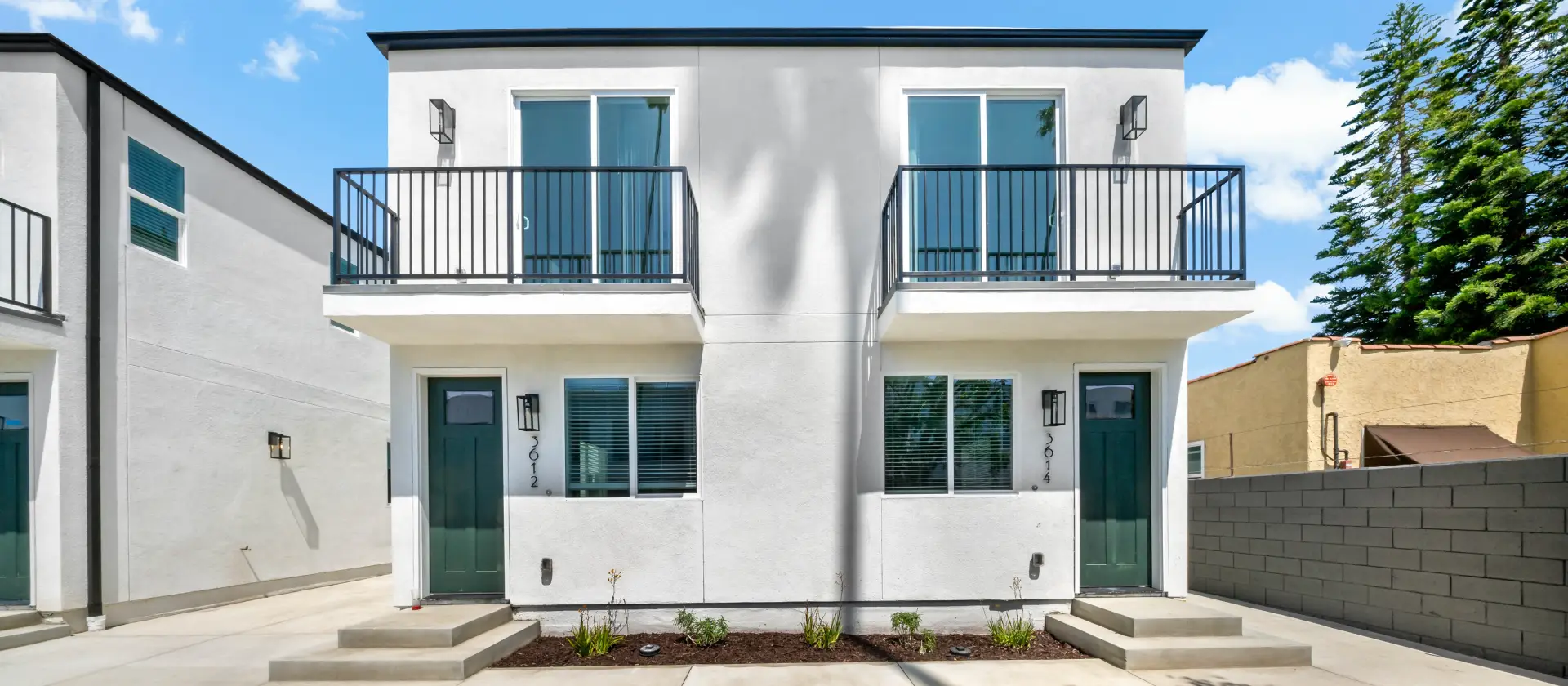 Front exterior view of 36TEN property with white facades, green doors, balconies, and a central walkway lined with palm trees.